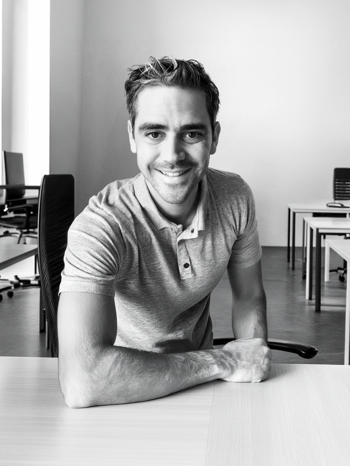 A smiling man in a gray polo shirt sits at a desk in a modern office space, showcasing a welcoming home office environment. - Alexi Hall