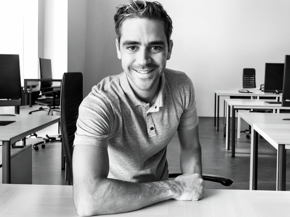 A smiling man in a gray polo shirt sits at a desk in a modern office space, showcasing a welcoming home office environment. - Alexi Hall
